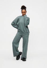 Πράσινο φαρδύ φούτερ και αντίστοιχο παντελόνι με φαρδιά μπατζάκια και μαλακή υφή. Το μοντέλο φοράει μαύρα chunky sneakers. Απλό λευκό φόντο.