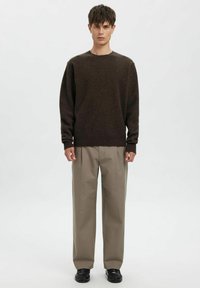 Rjavo pleten pulover, sproščen kroj, dolgi rokavi. Bež hlače po meri, ravne noge. Črni čevlji. Enobarvno belo ozadje.