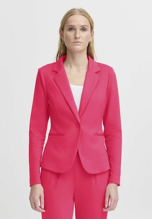 Donna con capelli biondi lunghi indossa un blazer rosa brillante su misura e pantaloni coordinati sopra una maglietta bianca, in piedi davanti a uno sfondo grigio uniforme.