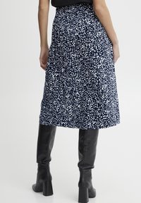 Žena oblečená v midi sukni s námořnickým modrým a bílým vzorem a černými vysokými podpatky po kolena, zobrazená zezadu na jednobodovém pozadí.