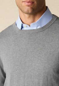 Szary sweter wełniany z okrągłym dekoltem nałożony na jasnoniebieską koszulę z kołnierzykiem. Tekstura wydaje się miękka z delikatnym, gładkim wykończeniem.