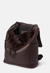 Mochila de cuero marrón con cierre de cordón, textura suave, bolsillo lateral y correa ajustable. Presenta un diseño minimalista.
