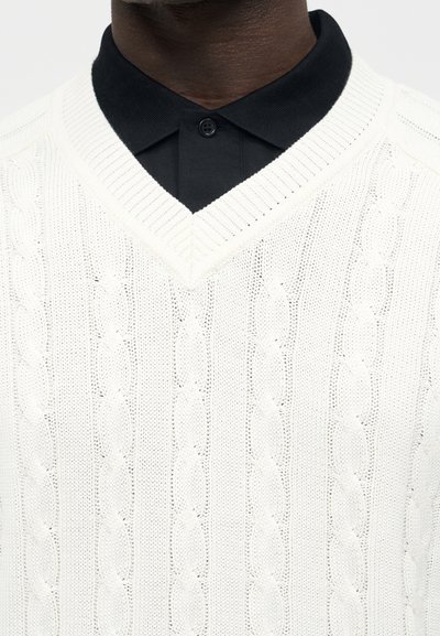 Άτομο με σκουρόχρωμο δέρμα που φοράει λευκό πουλόβερ με V-λαιμόκοψη και πλέξη με καλώδια πάνω από μαύρο πουκάμισο με γιακά και κουμπωμένο λαιμό.