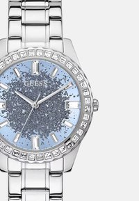 Srebrny zegarek Guess z okrągłą tarczą, b�łyszczącym niebieskim cyferblatem, kryształowymi detalami wokół obręczy i polerowaną metalową bransoletą.