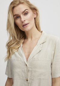 Chemise en lin beige clair à manches courtes avec un col en V, deux poches poitrine et une fermeture à boutons. Texture douce avec peu de plis.