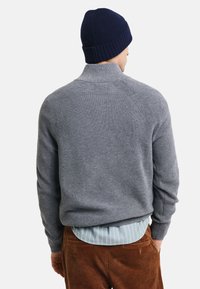 Grauer gerippter Pullover mit hohem Kragen, ausgestattet mit Raglanärmel. Kombiniert mit einem hellgestreiften Hemd und braunen Cordhosen. Marineblaues Strickmütze.