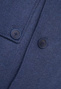 Marineblaues, strukturiertes Gewebe mit einem Fischgrätenmuster, das zwei dunkle Knöpfe auf einem Kleidungsstück zeigt und die Tiefe und Details des Stoffes betont.