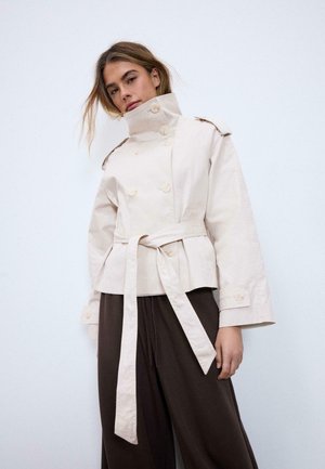 Ženska, oblečena v bež trenč plašč s pasom in visokim ovratnikom ter širokimi temno rjavimi hlačami, stoji pred belim ozadjem.