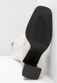 Botte blanche à talon allongée sur le côté, montrant une semelle noire texturée avec des lignes de traction diagonales et un talon carré bloc.