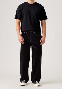 Homme portant un t-shirt noir à manches courtes, un pantalon large en velours côtelé noir, des baskets noires et blanches, debout devant un fond clair uni.