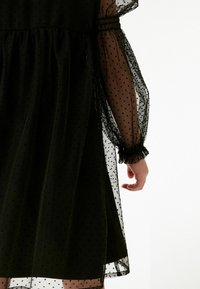 Schwarz gepunktetes Kleid mit durchsichtigen, puffartigen Ärmeln und geraffter Taille. Der Stoff hat ein weiches, strukturiertes Aussehen und eine fließende Silhouette.
