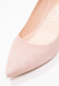 Escarpin à bout pointu en suède rose pâle, avec une texture lisse et un col ouvert. Minimaliste, sans hardware visible.