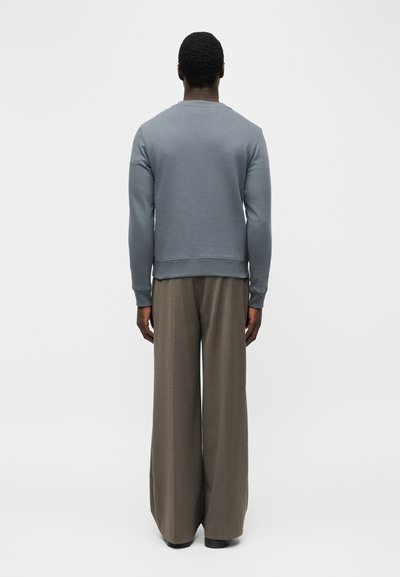 Άτομο που στέκεται με την πλάτη προς τα εμπρός, φοράει γκρι μακρυμάνικο πουκάμισο και χαλαρά καφέ παντελόνια, με λευκό απλό φόντο.