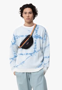 Světle modrý svetr s bílým tie-dye vzorem, kombinovaný s černou kabelkou přes rameno s vícebarevným pruhovaným popruhem.