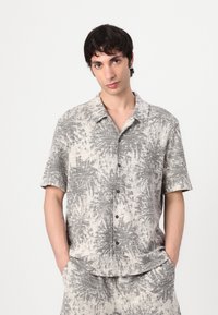 Camicia a maniche corte con bottoni di colore beige chiaro, decorata con un motivo astratto nero. Il tessuto sembra testurizzato, con una vestibilità rilassata e colletto aperto.