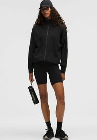 Čierna zipsová bunda s zakrivenými švami, skombinovaná s čiernymi cyklistickými šortkami. Model drží čiernu fľašu na vodu. Čierna čiapka a tenisky dokončujú vzhľad.