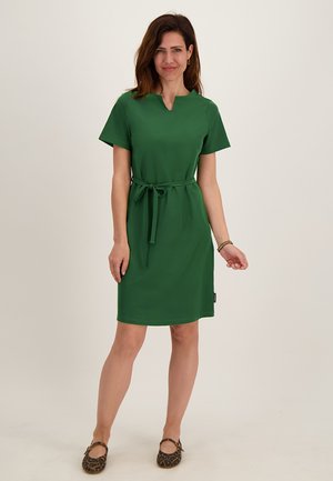 Vrouw staat in een knie-lange groene jurk met korte mouwen en een ceintuur in de taille, draagt leopardprint platte schoenen tegen een effen achtergrond.