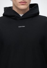 Άτομο που φοράει μαύρο φούτερ Calvin Klein με το όνομα της μάρκας με μικρά λευκά γράμματα στο στήθος, εμφανίζεται από τους ώμους ως το στόμα.