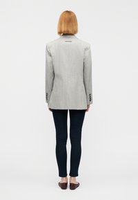 Blazer gris sur mesure avec une fente unique au dos, boutons noirs aux manches, associé à un jean slim foncé et des chaussures plates marron.