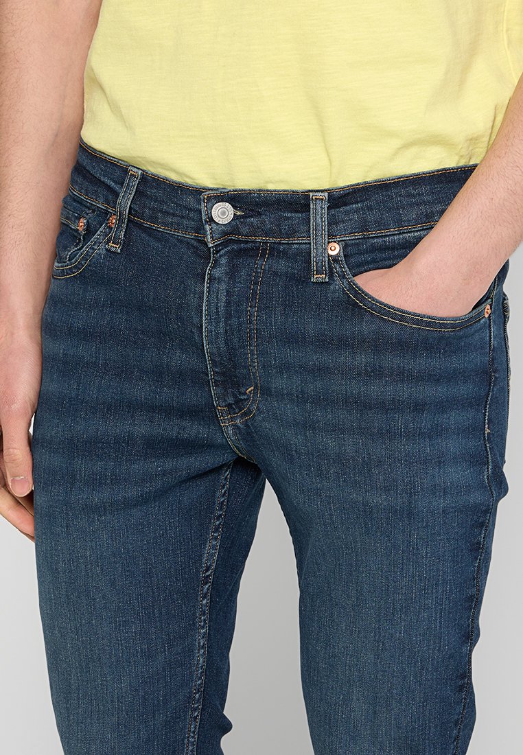Mann in dunkelblauen Jeans und einem hellgelben Hemd, mit der rechten Hand in der Jeanstasche und dem linken Arm entspannt an der Seite.