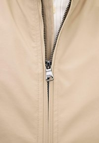 Nærbillede af en beige jakke, hvor lynlåsen er delvist trukket op, med metal-lynlåstræk og synlige tænder på stoffet.