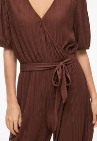 Rudas, plisuotas kombinezonas su giliu V formos iškirpimu, trumpomis pūstomis rankovėmis ir savarankišku diržu. Minkšta tekstūra pabrėžia struktūrizuotą dizainą.