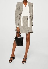 Blazer gris a cuadros sobre una blusa blanca, combinado con una falda a juego, un bolso tipo cubo negro y sandalias negras de tacón con dos tiras.