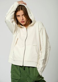 Sudadera cremosa con cremallera hecha de un tejido suave, que presenta tachuelas doradas a lo largo de las mangas, una capucha y bolsillos frontales. Combinada con pantalones verdes.
