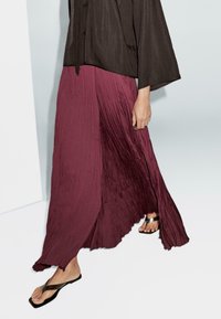 Jupe maxi plissée prune avec un design fluide, associée à un haut brun ample et des tongs noires, mettant en valeur une texture lisse et le mouvement.