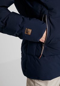 Tmavě modrá prošívaná bunda s kapsou na zip, hnědým logem na rukávu a texturovanou látkou s nadýchaným designem.