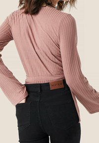 Ljusrosa ribbad långärmad topp med en knytning i midjan, kombinerad med svarta högmidjade jeans med en läderlapp märkta "NA-KD."