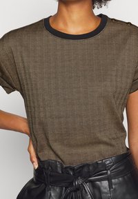 Brun mönstrad kortärmad skjorta med ett fiskbensmönster och svart ribbat halsband, bärs med höga svarta läderbyxor.