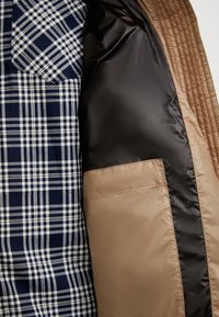 Binnenkant van een bruine gewatteerde jas die een marineblauw en wit geruit overhemd met een borstzakje toont, met een gladde zwarte voering en een beige stofaccent.
