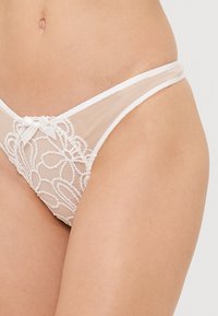 Γυναικείο στρινγκ από γυμνό διχτυωτό ύφασμα με λευκή δαντέλα, διακοσμημένο με λουλουδάτο σχέδιο, featuring μια μικρή κορδέλα στο μπροστινό μέρος και λεπτές, αντίθετες άκρες στη μέση.