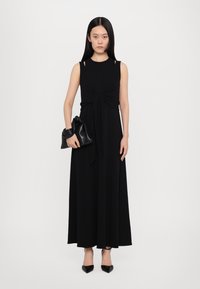 Svart ermeløs maxikjole med knyttet frontdetalj, glatt stoff og to små utskjæringer på skuldrene. Kombinert med en svart clutch.