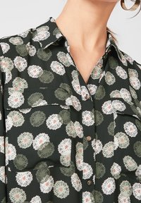 Maglietta nera con motivo floreale in verde, bianco e rosa. Include due tasche sul petto e bottoni metallici. Tessuto morbido.