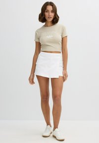 Camiseta corta beige con texto blanco "BREATHING", combinada con una falda envolvente blanca. La modelo lleva zapatillas blancas con suelas texturizadas.
