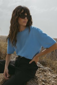 Gaiši zilā krāsā īsajām piedurknēm svīteris mīkstā adīšanas audumā, saskaņots ar augsto jostasvietu melnajām biksēm. Modelis valkā pārmērīgi lielas melnas saulesbrilles.