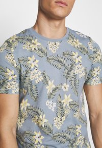 Modré tričko s krátkým rukávem s květinovým vzorem se žlutými květy a zelenými listy, vyrobené z měkké bavlny.