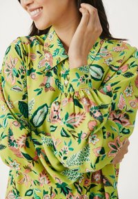 Blouse florale verte avec un col, des manches longues avec des poignets froncés, et un motif vibrant de feuilles et fleurs roses et vertes foncées.