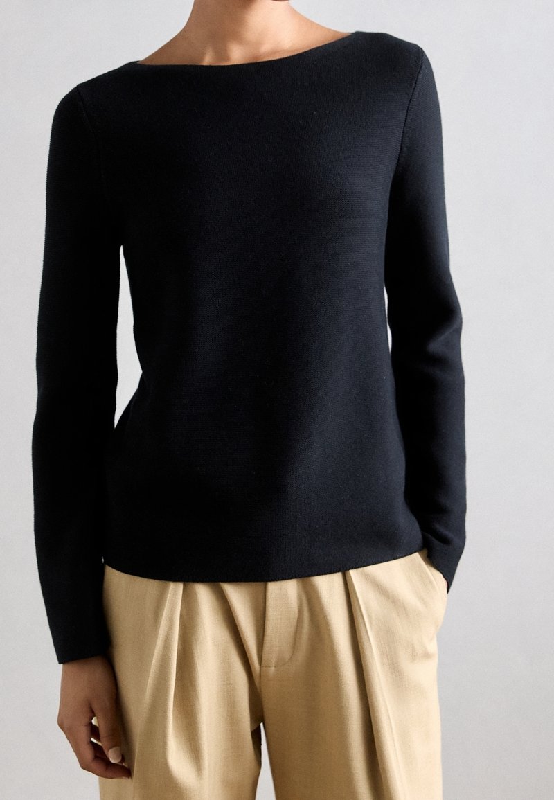 Personne portant un haut noir ajusté à manches longues et un pantalon beige plissé taille haute avec des mains dans les poches sur un fond uni.