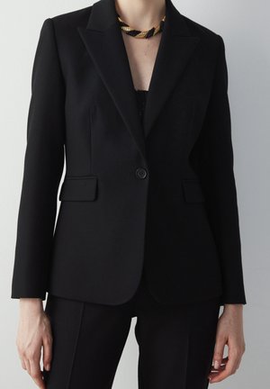 Blazer noir ajusté avec épaules structurées, fermeture à un bouton et deux poches avant. Tissu lisse et coupe ajustée, associé à un pantalon noir.