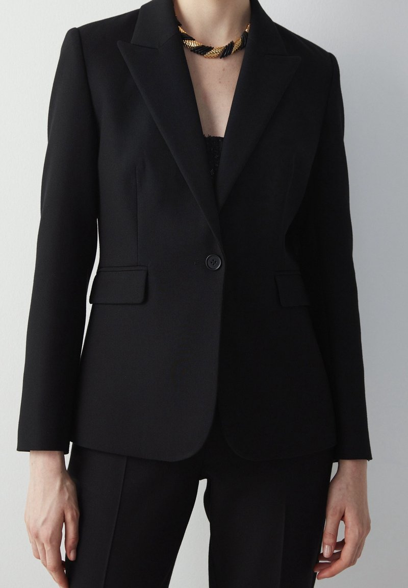 Černý přizpůsobený blazer se strukturovanými rameny, zapínáním na jeden knoflík a dvěma předními kapsami. Hladká látka a přizpůsobený střih doplněný černými kalhotami.