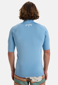 Kortærmet blå rash guard lavet af glat stof, med et hvidt logo på ryggen og minimal sømdetaljering.