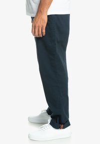 Pantalon en coton bleu marine à coupe droite, avec un petit logo et des poignets retroussés, associé à des baskets blanches.