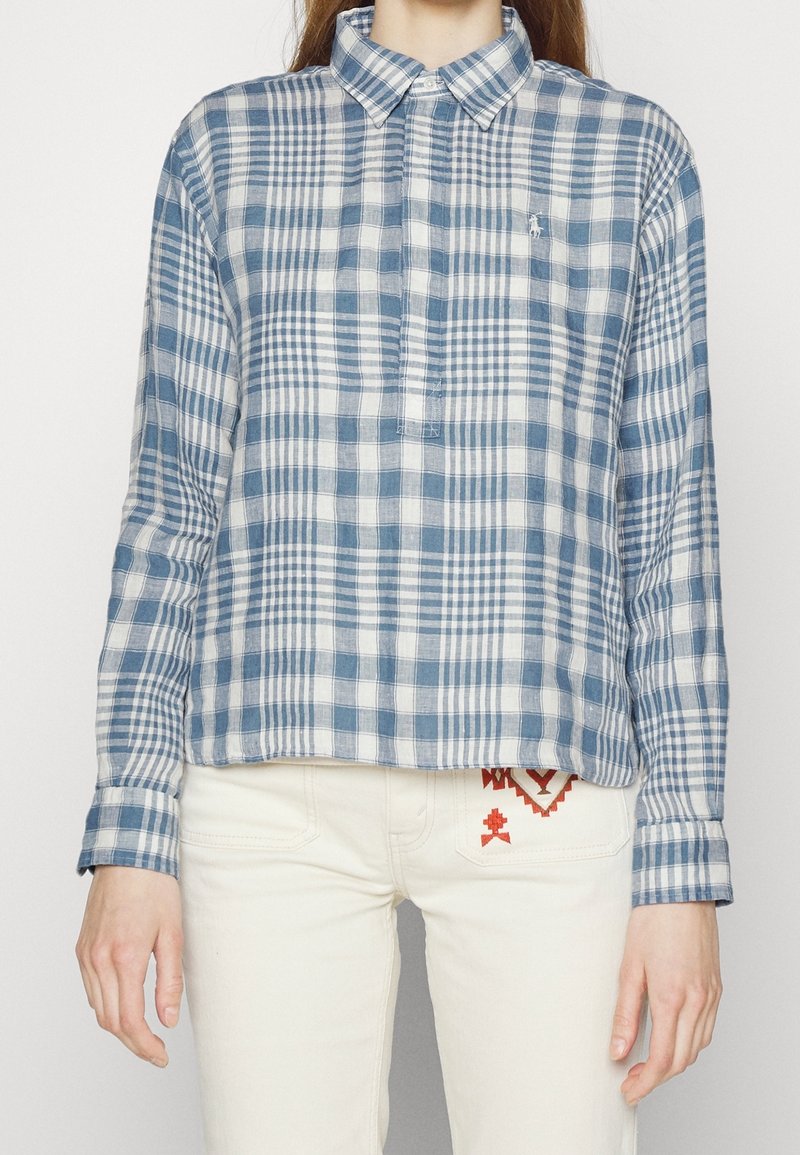 Personne portant une chemise à carreaux bleue et blanche boutonnée avec un pantalon blanc présentant des motifs brodés rouges sur la poche.
