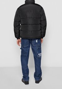 Czarna kurtka puchowa o pikowanej teksturze i wysokim kołnierzu, zestawiona z niebieskimi dżinsowymi spodniami typu cargo z kieszeniami i logo.