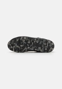 Zapato negro de exterior con suela texturizada que presenta un patrón de camuflaje en gris y negro; diseñado para ofrecer tracción en superficies variadas.