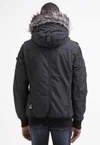 Giacca nera imbottita con cappuccio foderato in pelliccia sintetica, polsini a costine e molteplici tasche pratiche. Presenta dettagli cuciti e un'etichetta con logo.