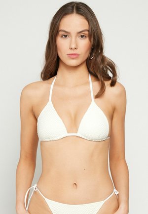 Młoda kobieta o falowanych brązowych włosach, ubrana w teksturowany biały trójkątny top od bikini i pasujące do niego majtki, stojąca na jednolitym tle.